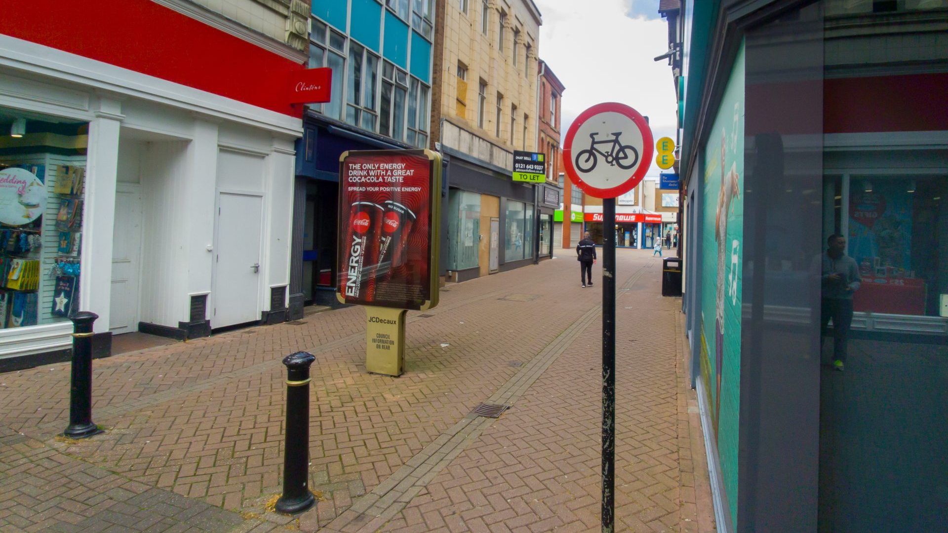 A no-cycling sign on an upright pole at the entrance to a narrow pedestrianised town centre street.