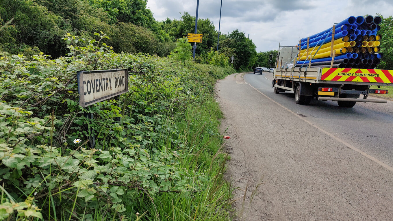 Shared use path with foliage to the left and a road name sign labelled "Coventry Road". To the right of the path is the main carriageway when a truck loaded with pipework has just gone past.
