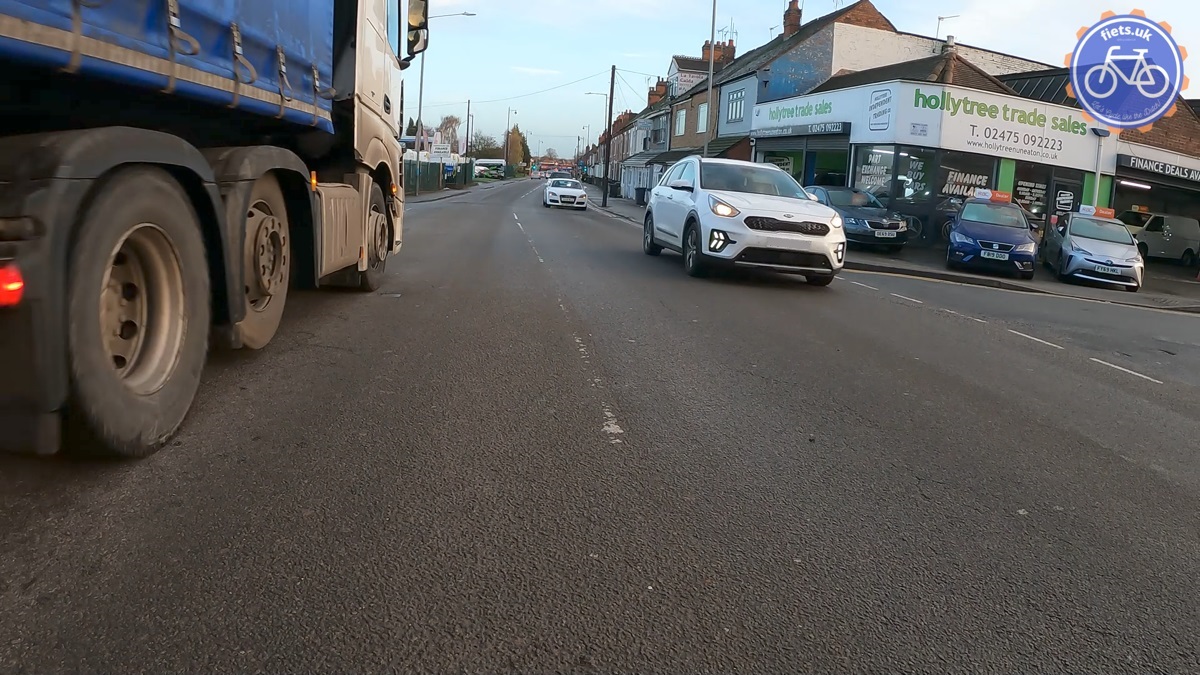 Cycle camera view of a main urban road, waiting to turn right to a smaller road. An HGV passes on the left (nearside) as traffic approaches in the opposite direction preventing the turn.