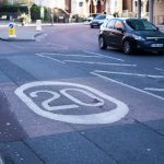 A 20mph roundel painted on an urban road surface just before the road bends round to the right. A car travels in the opposite direction. Image is by Tom Page (https://flic.kr/p/s9tMmY). Creative Commons Licence BY-SA 2.0.