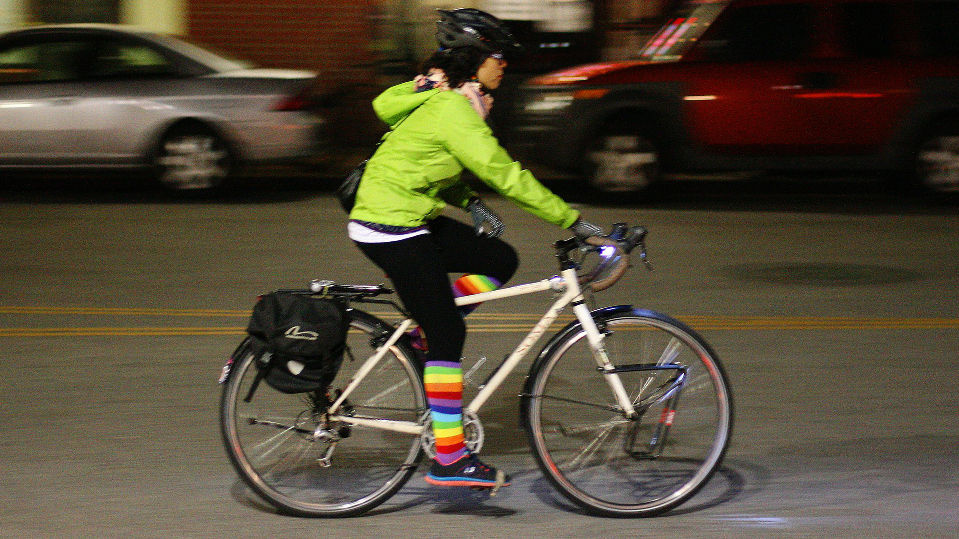 Person riding a bicycle at night, dressed in a high-visibility long-sleeved jacket and rainbow-striped socks. Image by Richard Masoner / Cyclelicious (Creative Commons CC BY-SA 2.0 DEED) - https://flic.kr/p/qHkNvC
