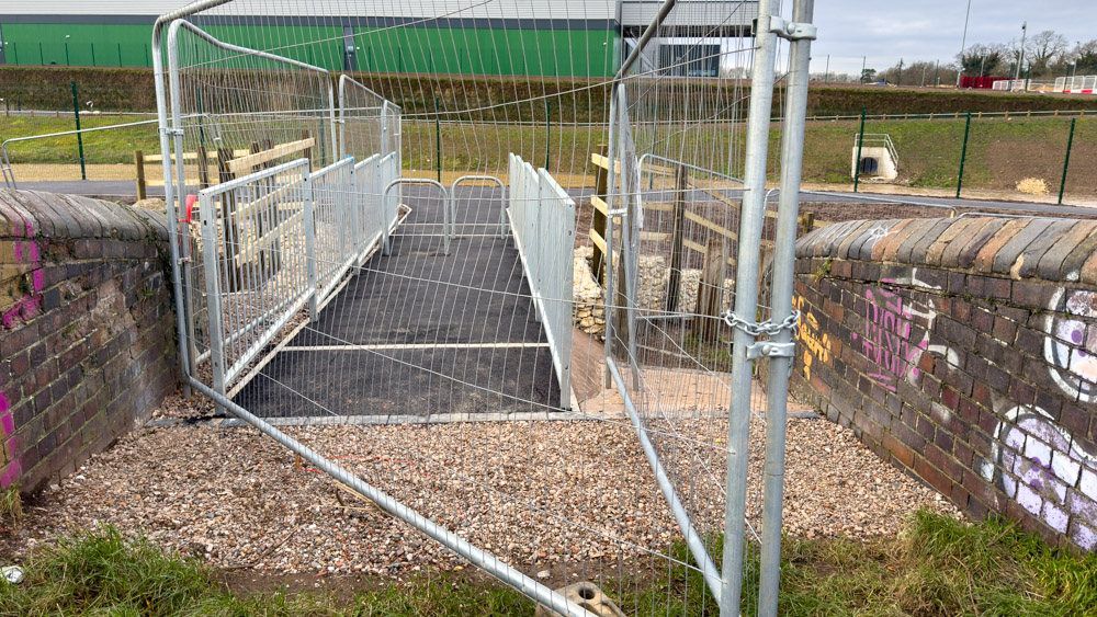 A fenced off short stub of pathway between an old canal bridge and a main path. The stub is about 1.6m wide and flanked with metal railings. Two barriers are in the path, arranged in a chicane style.