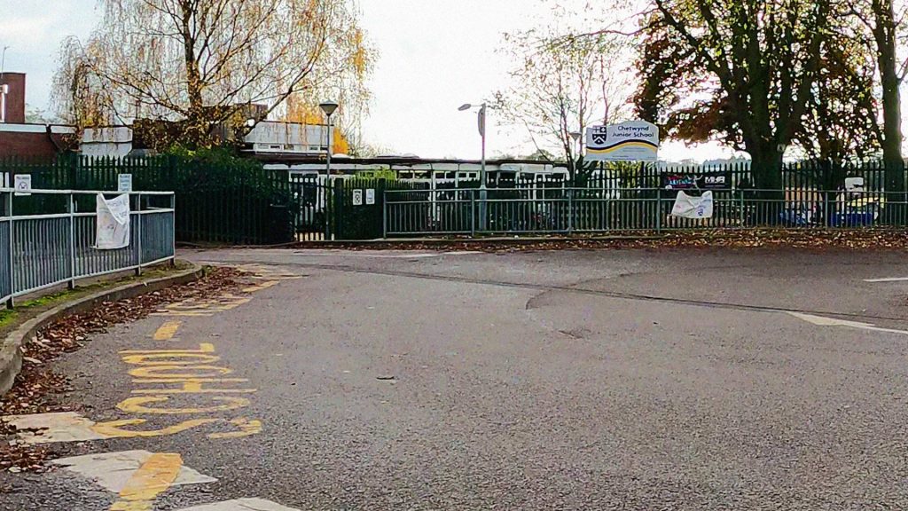 A primary school seen from the public road. The road joins an area for a mini-roundabout, the centre of which is just out of shot to the right. "School Keep Clear" zig-zags surround the boundary of the road. The footpath is buffered to the road by guard rails. The school's sign can be seen standing high, above the school's green fencing.