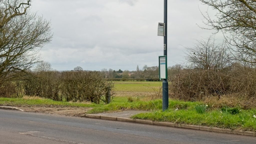 A bus stop on the far side of the carriageway with a small amount of hard-standing but otherwise no pavement; just a grass verge and hedgerow. There is no shelter or seating. Just a pole with a bus stop sign and timetable board. Beyond is a field which will eventually be built over.