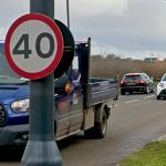 A 40mph speed limit sign beside a road with a flat-bed van heading towards the camera, followed by a small white car. Another car is travelling in the opposite direction. The far side of the two-lane road is bounded by hedgerow and a field; there is no pavement.