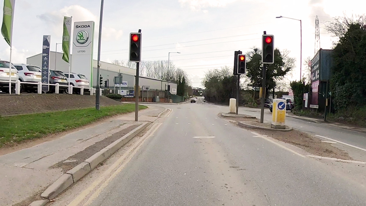 Longford Road southbound, approaching red traffic signals. In the distance, the road runs under a motorway bridge. A Skoda car showroom can be seen to the left.