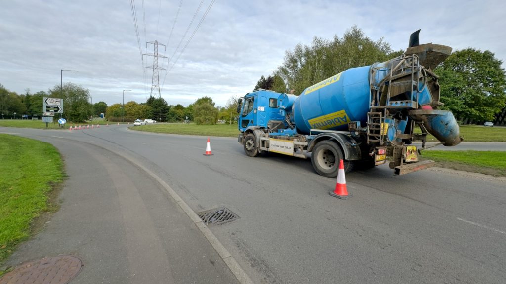 A mixing truck waits to join the Longford Road roundabout. Powerlines cross overhead. Cones have closed off the left hand lane.