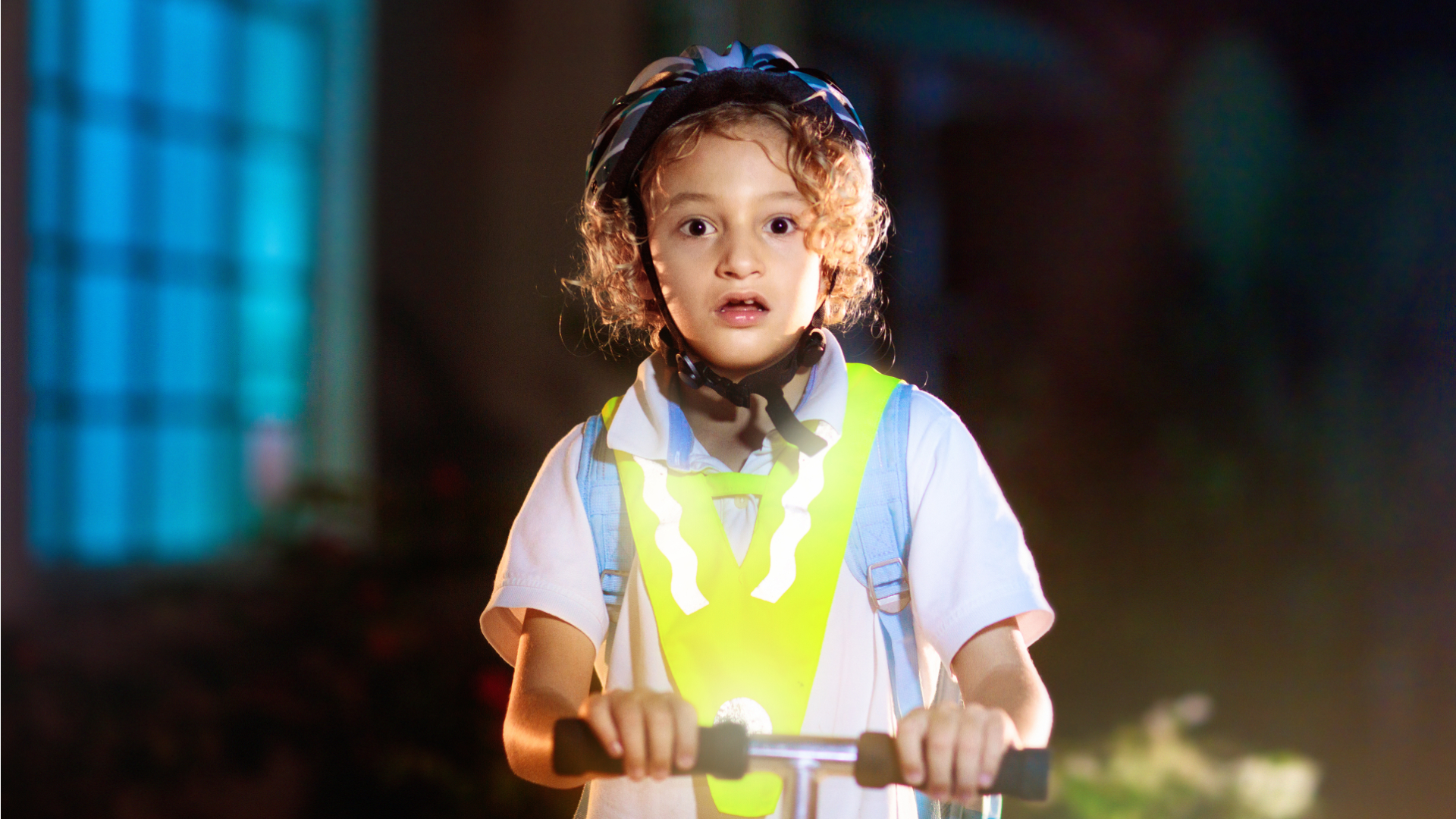 Child in a reflective vest at night, holding the handlebar of a scooter, with a slightly startled or worried look on their face.